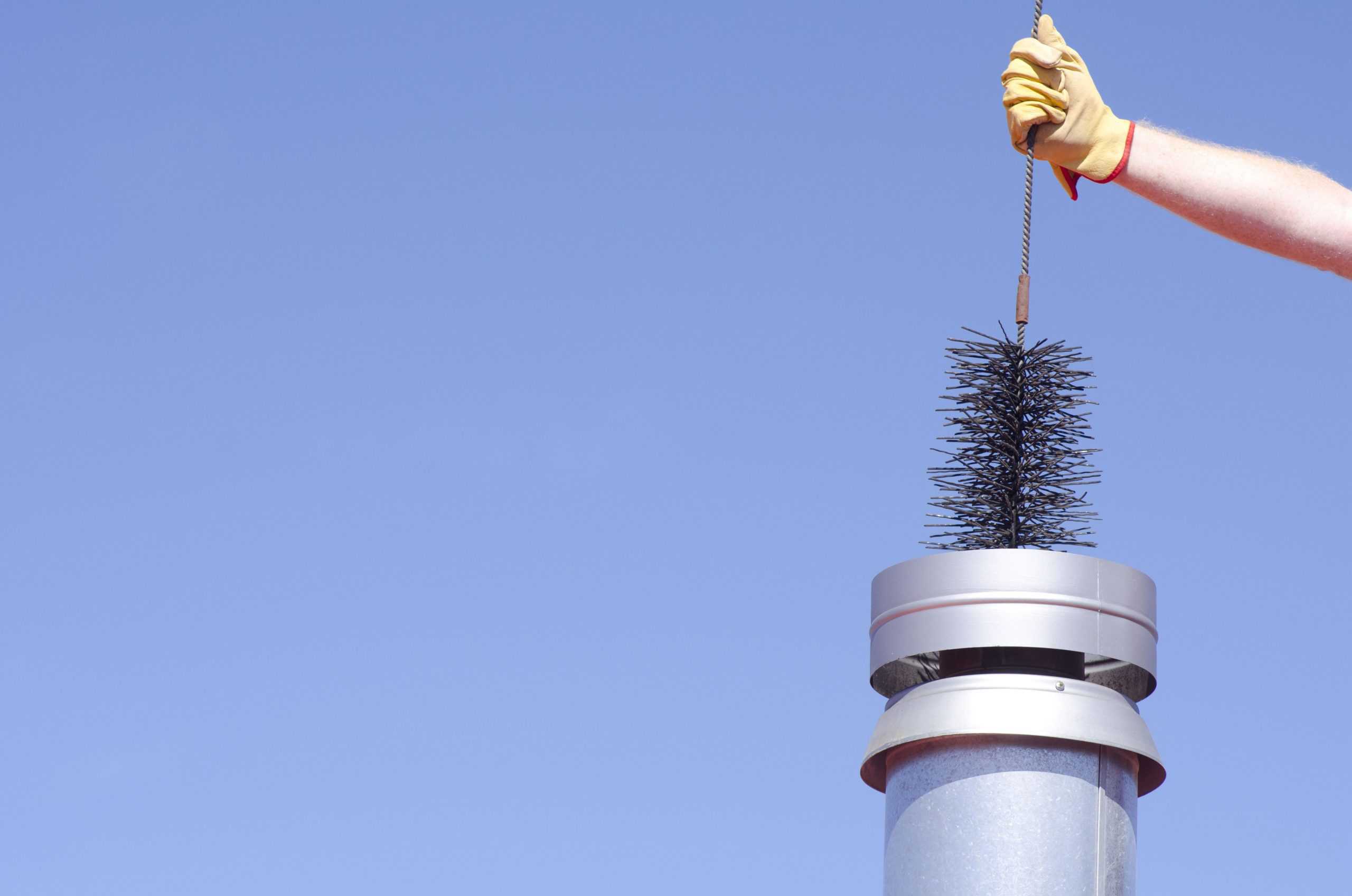 A person wearing a yellow glove is holding a chimney sweep brush above a metal chimney pipe, demonstrating expert chimney cleaning skills against the backdrop of a clear blue sky.