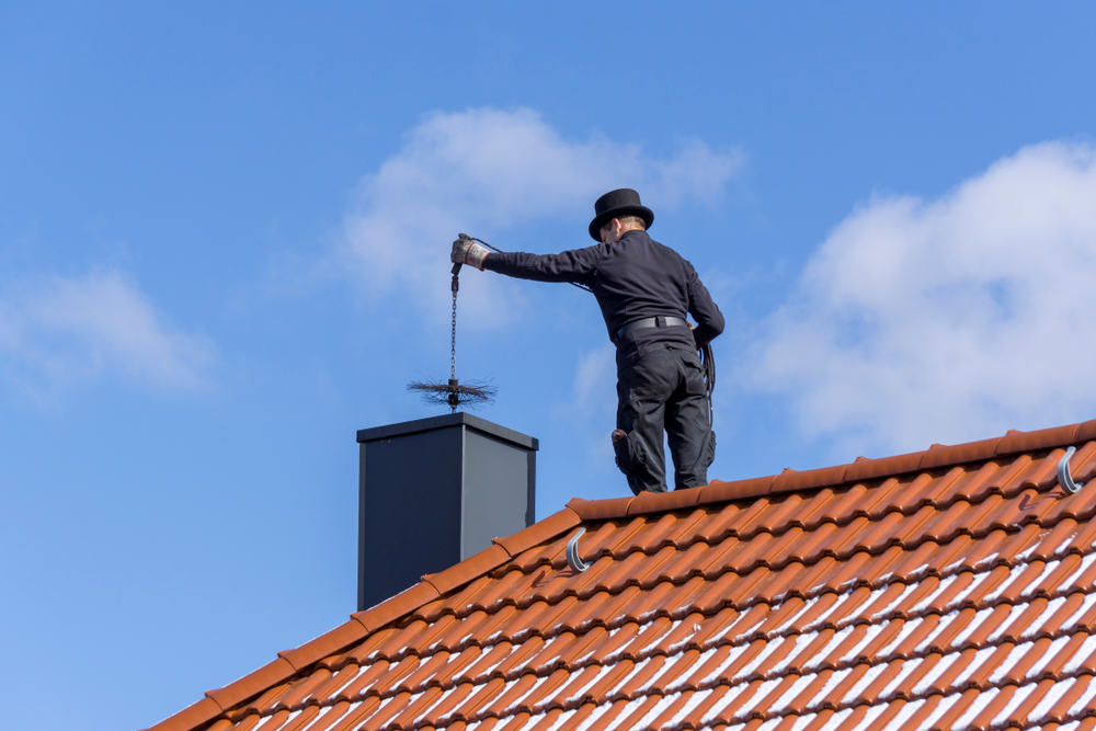A chimney sweep in a black uniform and top hat expertly performs chimney cleaning on a red-tiled roof under a blue sky with a few clouds.
