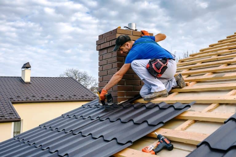 A worker in a blue shirt and gloves installs black roofing tiles on a house. Kneeling near the brick chimney, surrounded by tools, he prepares to fit chimney caps under a partly cloudy sky.