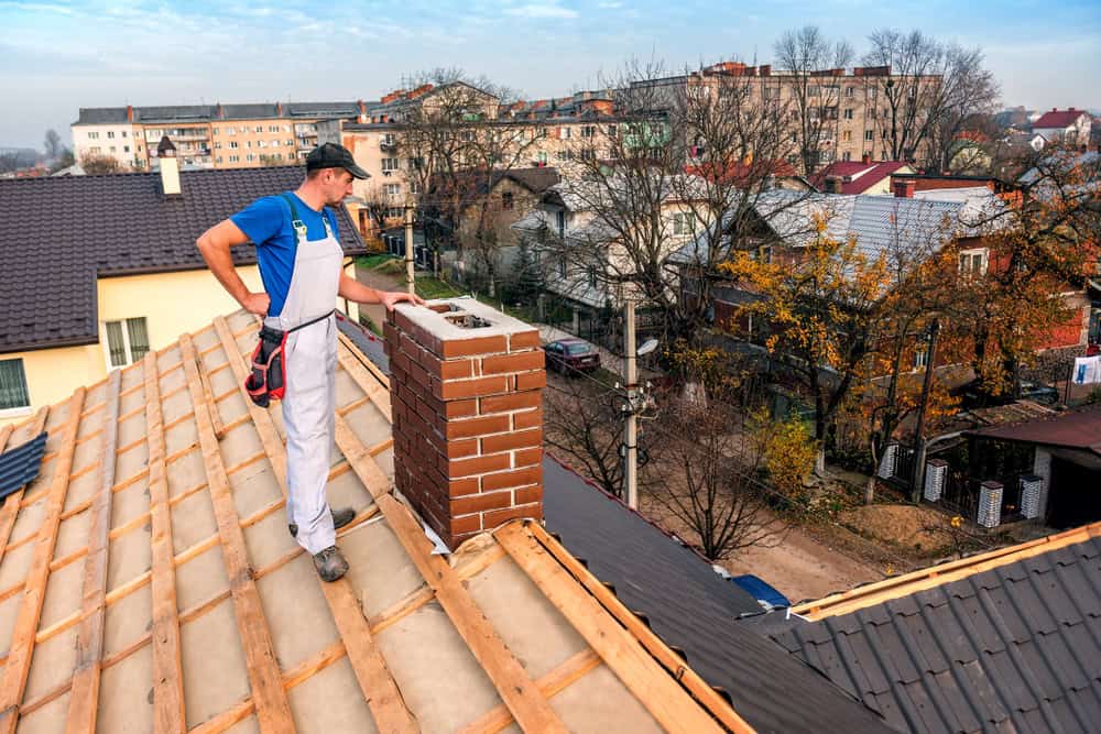 A construction worker in a blue shirt and white overalls stands on a tan shingle roof conducting a chimney repair. The background shows a residential neighborhood with trees and houses on an overcast day.