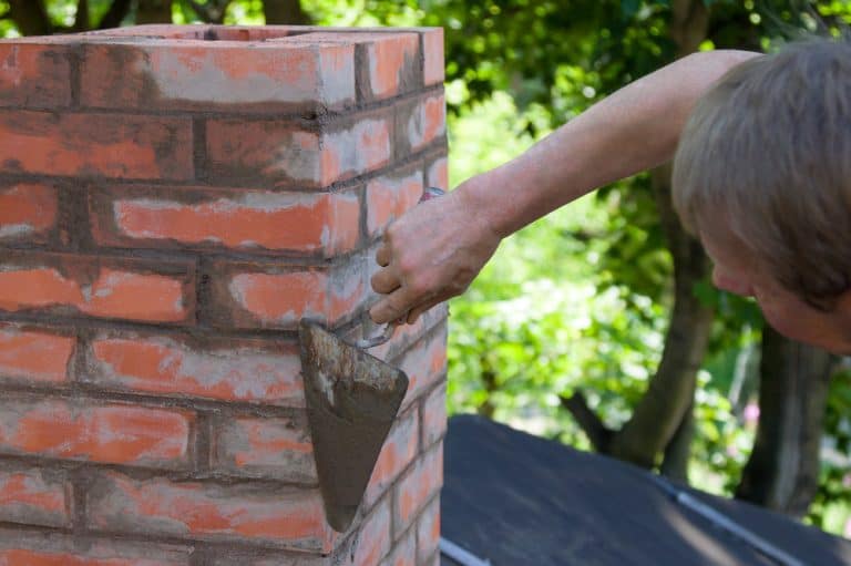 A person from a chimney company expertly uses a trowel to apply mortar to a red brick chimney. The outdoor scene is framed by lush green foliage in the background, enhancing the craftsmanship on display.