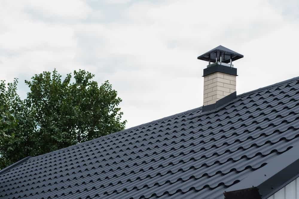 A close-up of a residential roof showcases dark metal tiles, while a beige brick chimney with a black cap hints at the expertise of a skilled chimney sweep. In the background, green tree leaves contrast against the cloudy sky.