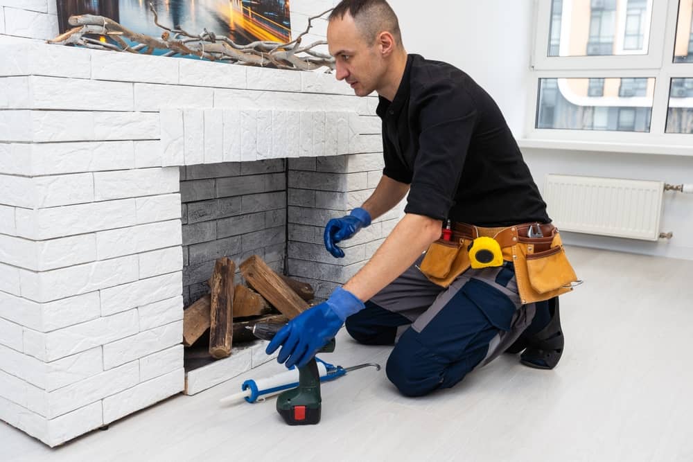A person kneels in front of a white brick fireplace, arranging logs. They are wearing blue gloves and a tool belt, with various tools from the chimney repair company scattered nearby. A large window is in the background.