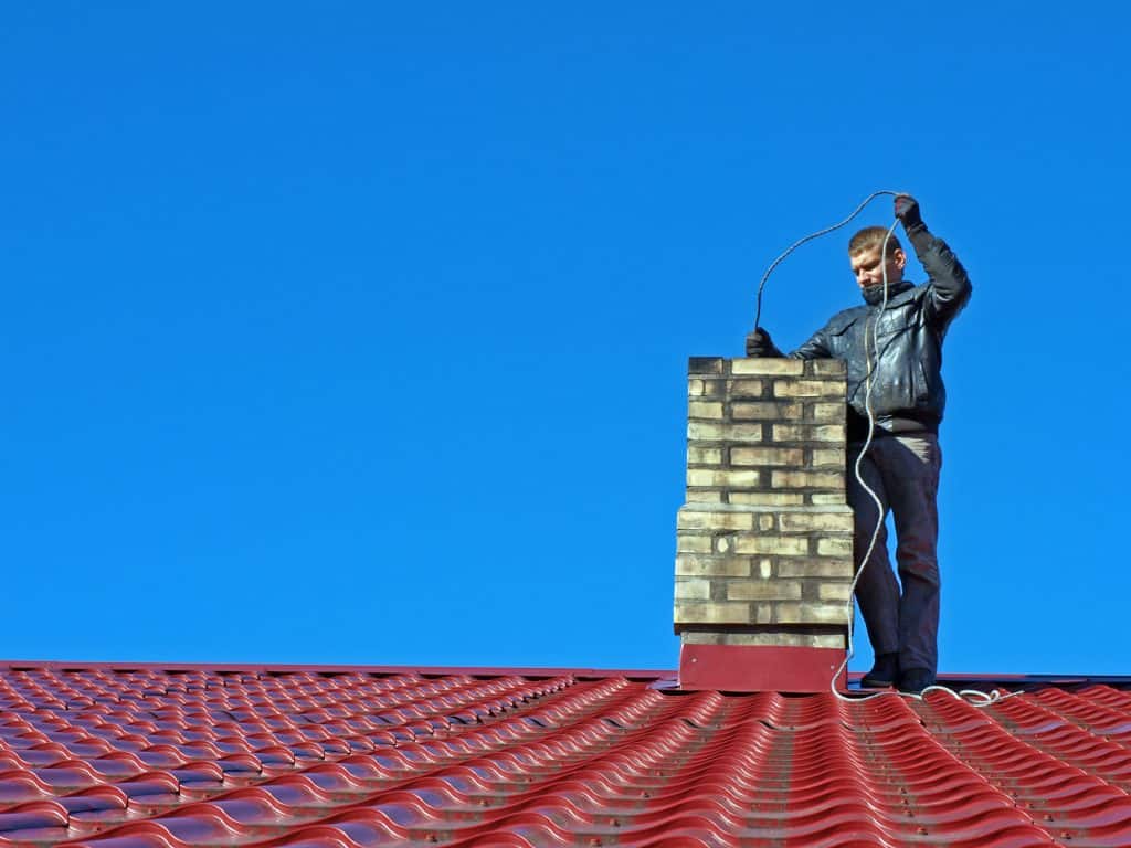 A person stands on a red corrugated metal roof under a clear blue sky, holding a long cable near the brick chimney. They're clad in a black jacket and dark pants, clearly busy with some chimney cleaning.