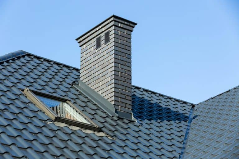 A sloped roof with blue-gray shingles features a pristine brick chimney, freshly tended to by a top-notch chimney cleaning service, and a skylight window, set against a clear blue sky.