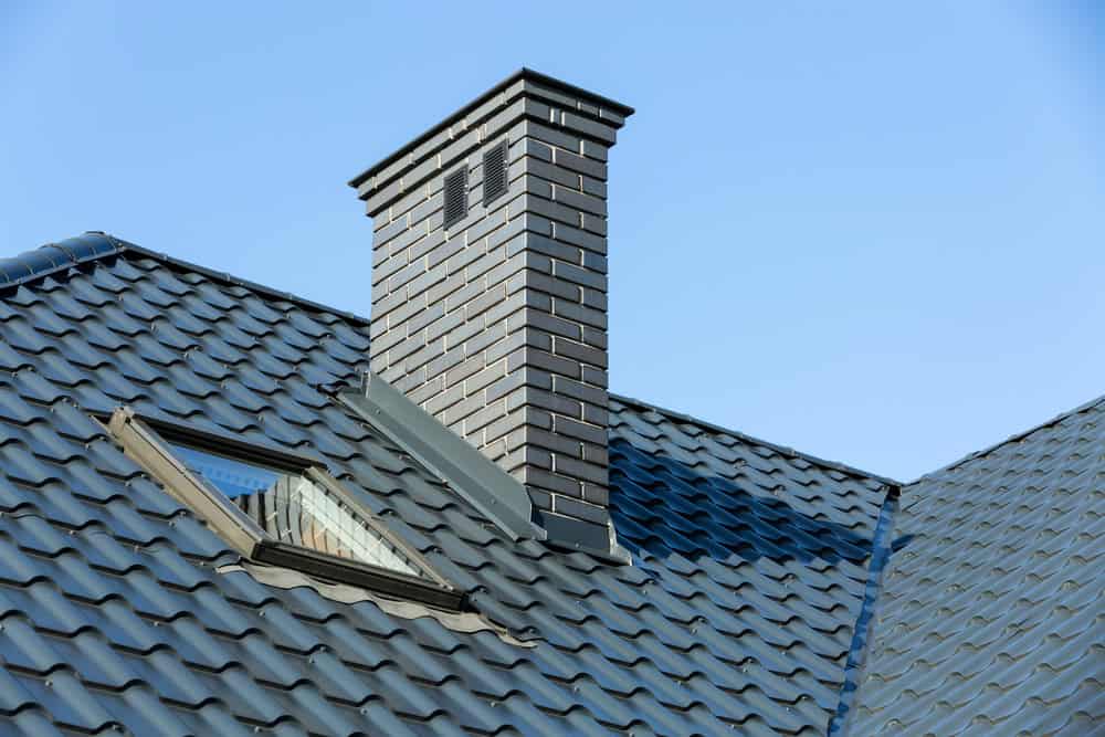 A sloped roof with blue-gray shingles features a pristine brick chimney, freshly tended to by a top-notch chimney cleaning service, and a skylight window, set against a clear blue sky.