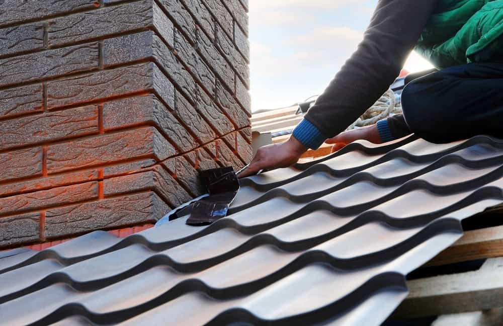A person installing metal roofing near a brick chimney carefully aligns the corrugated sheets for a proper fit, ensuring every detail complements the masonry. They're likely from a chimney company, given their precision and expertise, while the sky peeks through in the background.