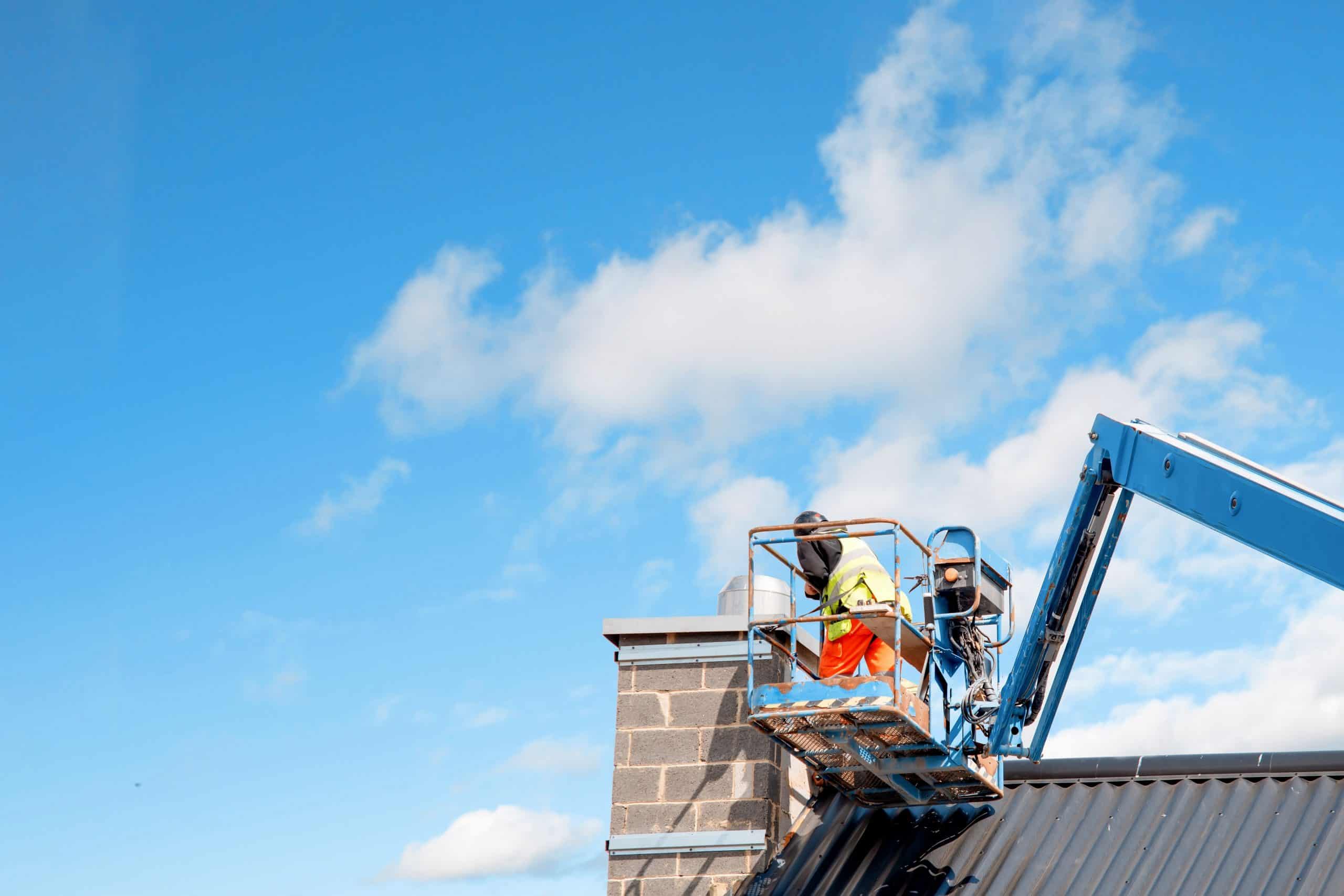 A worker stands on a blue lift, expertly performing chimney repair against a clear blue sky with scattered clouds. Wearing a neon safety vest and orange pants, they focus intently on the task. The lift is positioned beside the building's sloped roof.