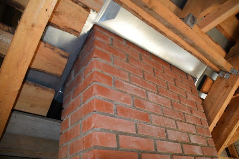 View of a brick chimney extending through a wooden roof structure under construction, showcasing exposed beams and insulation. A Chimney Company is installing features to enhance the setup.
