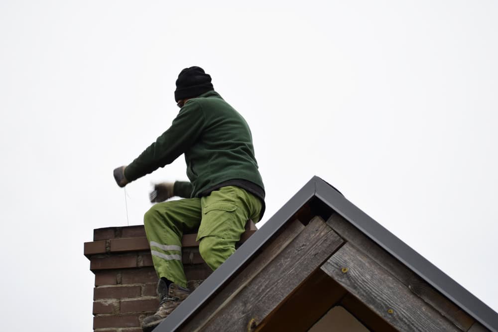 A person wearing a green jacket and pants is busy with chimney repair on a brick chimney. They sit on a rooftop, facing away from the camera, against a cloudy sky background.