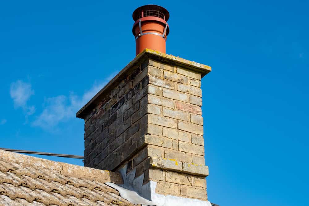 A brick chimney with a red metal cap sits atop a sloped roof, its design reflecting the professional touch of chimney repair. All this is set against a clear blue sky.