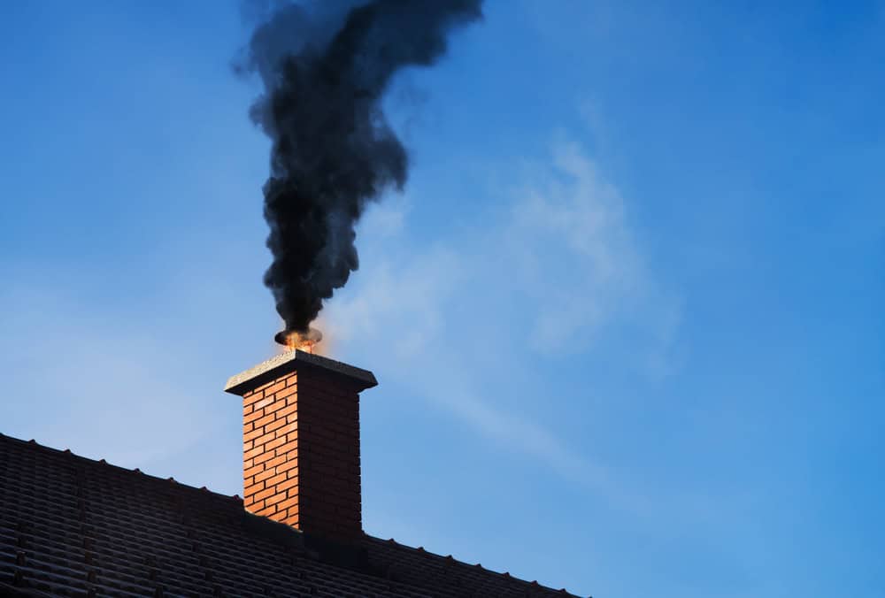 A brick chimney on a rooftop, expertly maintained by a renowned Chimney Company, emits thick black smoke against a clear blue sky.