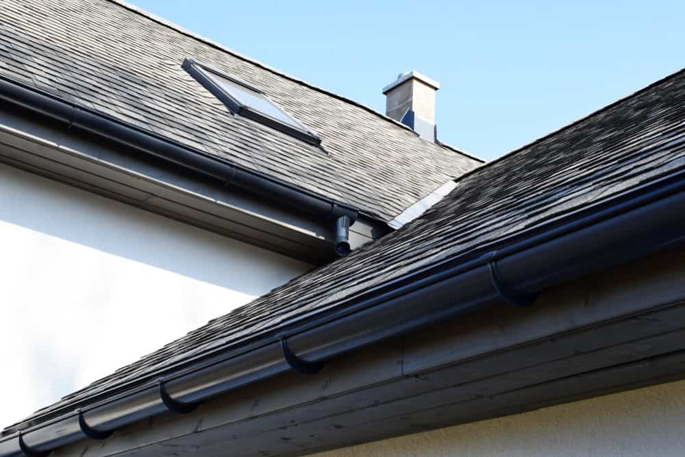 Close-up view of a house roof with dark shingles and a skylight. A chimney, possibly serviced by a local chimney repair company, is visible in the background. Black gutters run along the roof's edge against a clear blue sky.