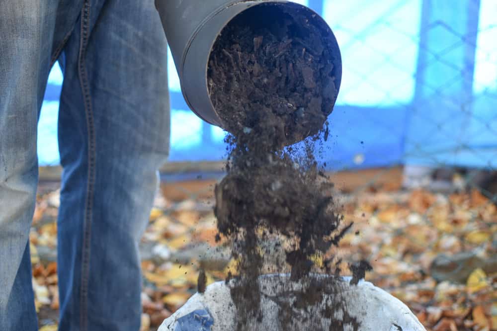A person in jeans empties dark soil from a black pipe into a container, much like the precision needed in chimney sweep tasks. Fallen leaves are scattered on the ground in the background.