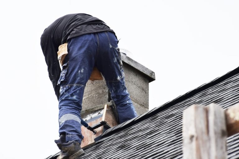 A person wearing work clothes and boots is standing on a sloped shingle roof, carefully inspecting the rectangular chimney. With wooden tools or materials nearby, they seem focused on installing chimney caps under the overcast sky. The person's back faces the camera.