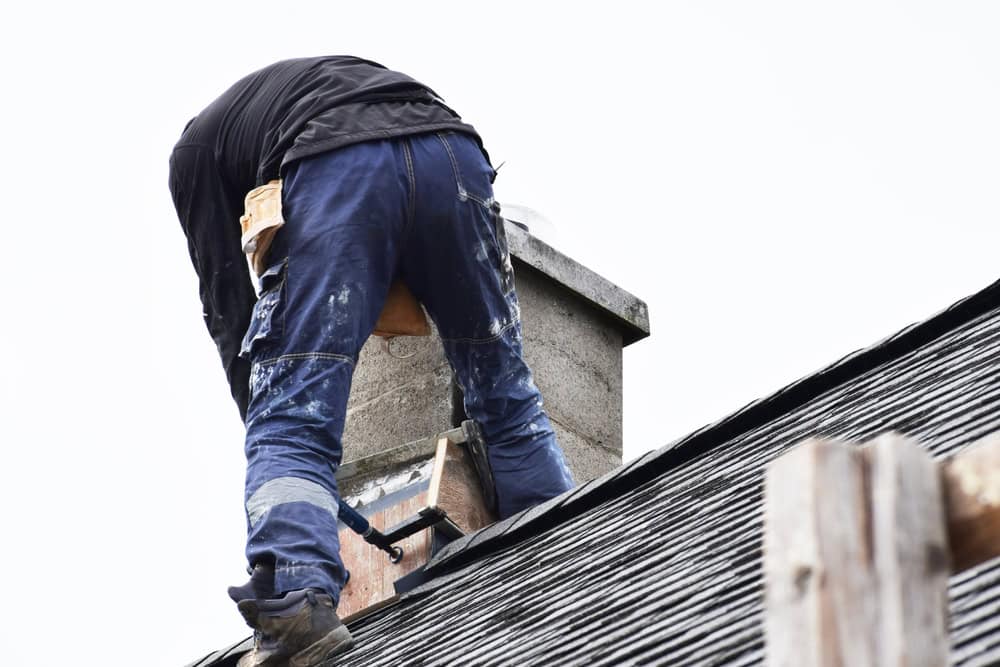 A person is diligently working on a rooftop next to a chimney, providing expert chimney services in Westchester County, NY. Clad in a black shirt, blue jeans with paint splatters, and sturdy boots, they focus on repairs with tools in a pouch under an overcast sky.