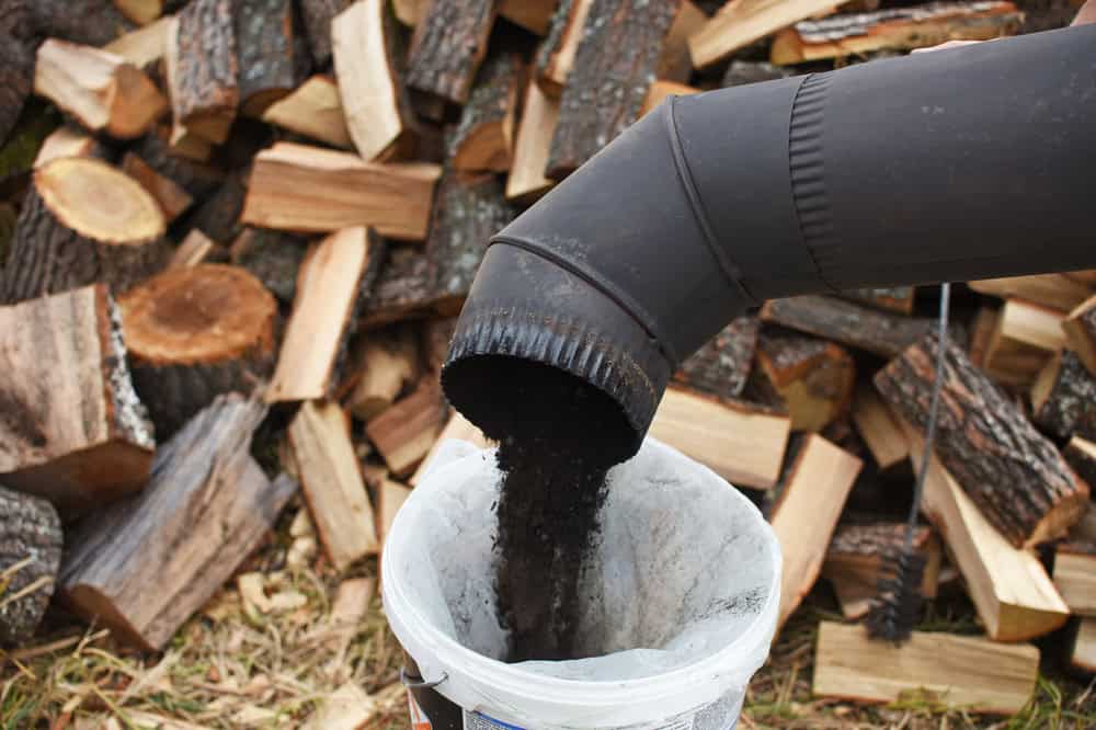 A chimney sweep's tool releases soot into a bucket, set against a backdrop of stacked wood logs. The dark soot pours steadily from a black, angled pipe.