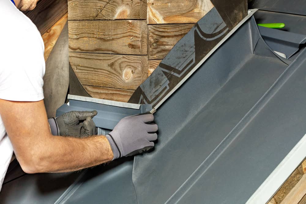 A person wearing gloves is installing metal flashing on a roof, carefully fitting the pieces together around a wooden structure during a meticulous chimney repair.