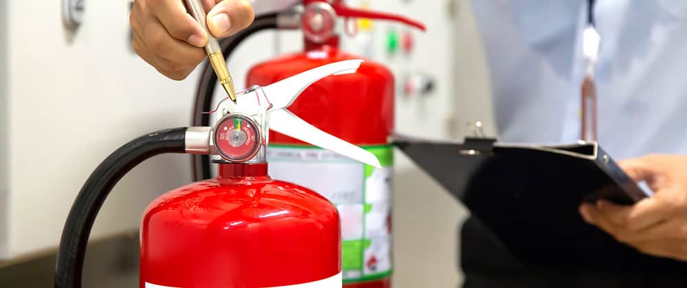 A person uses a pen to point at the gauge of a red fire extinguisher while holding a clipboard, much like a chimney sweep inspecting fireplace safety. Another fire extinguisher is visible in the background.