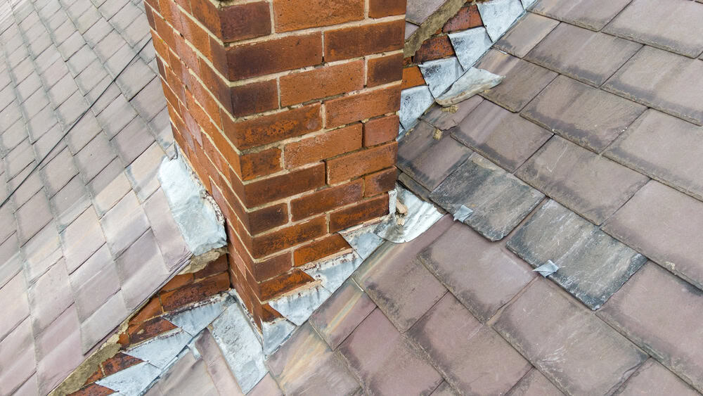 A brick chimney in need of cleaning stands surrounded by worn, weathered flashing and shingles. The discolored, uneven shingles hint at age and potential water damage, suggesting a possible need for chimney repair to prevent further issues.