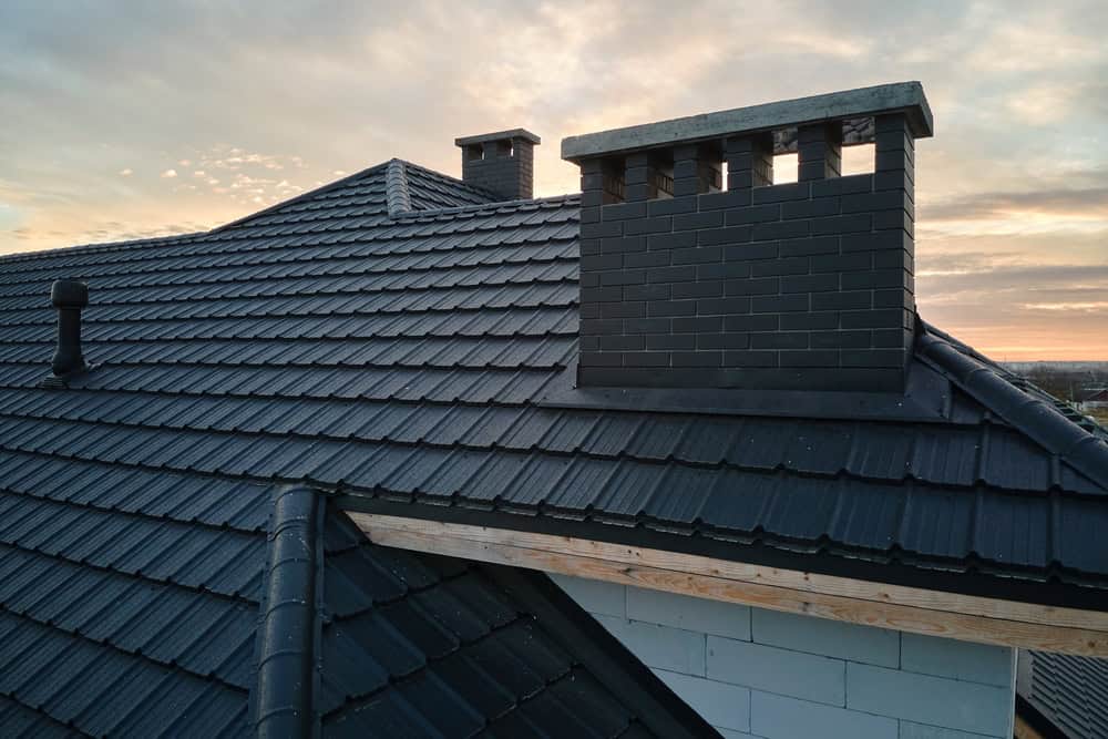 A rooftop with dark blue tiles and a brick chimney, recently serviced by a skilled chimney sweep, is shown at sunset. The sky is partly cloudy with a soft orange glow, and the roof features a wooden fascia board along the edge.