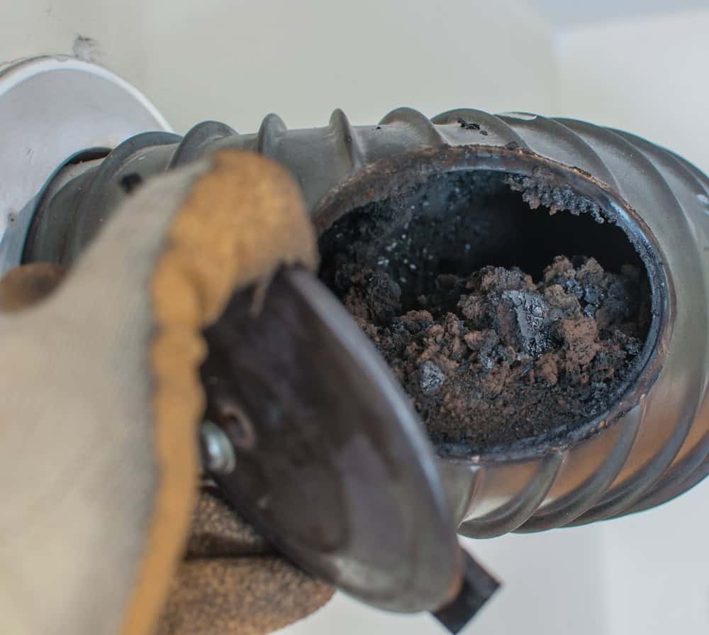 A gloved hand carefully opens a metal pipe, revealing an accumulation of dirt and debris inside. The pipe seems to be part of a ventilation system, hinting at the need for chimney repair by a professional chimney company.