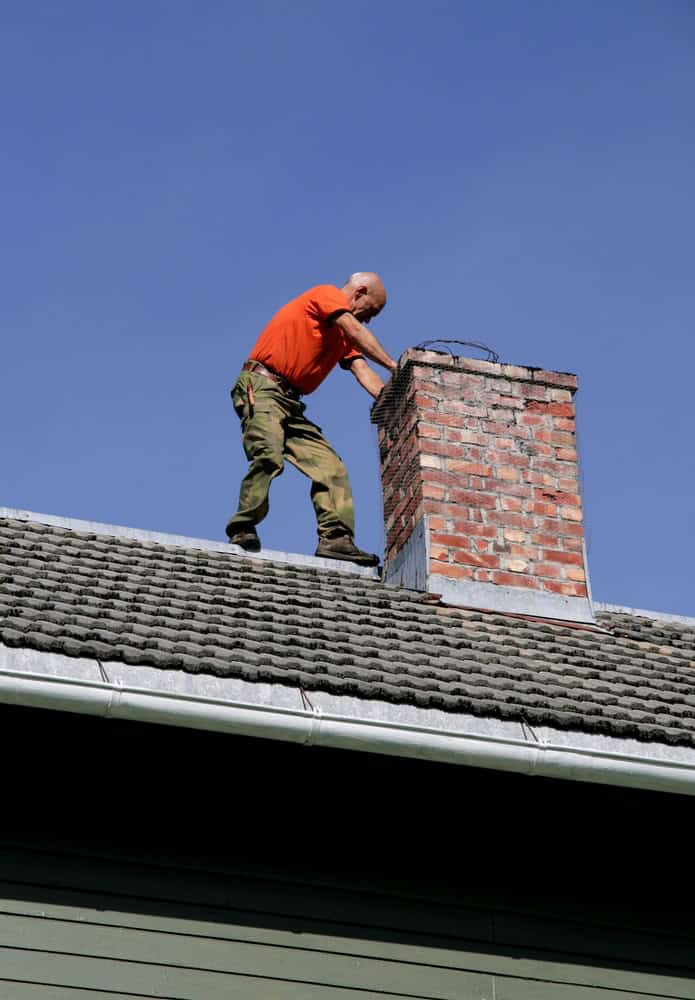 A person in an orange shirt and green pants stands on a roof next to a brick chimney, possibly involved in chimney repair. The clear blue sky provides a serene backdrop as they inspect or work on the chimney.
