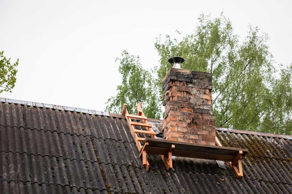 A weathered brick chimney stands on a corrugated rooftop, surrounded by green trees. A wooden ladder rests against the roof, leading to a small wooden platform with chimney caps surrounding it. The sky is overcast, setting the scene for scheduled maintenance by the Chimney Company.