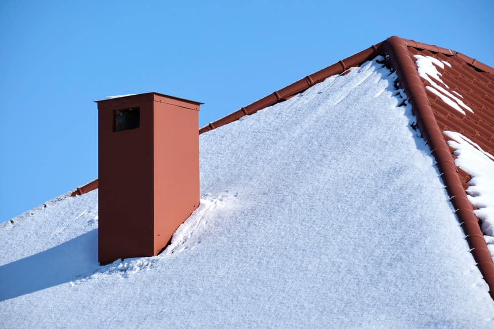 A red chimney, expertly maintained by a local chimney sweep, stands on a snow-covered roof under a clear blue sky. The snow forms a smooth blanket across the roof tiles.