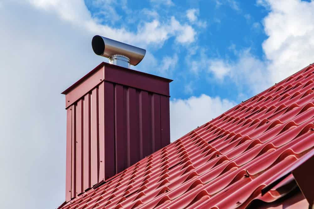 A red metal roof features a chimney topped by a cylindrical metal vent, expertly installed by a renowned Chimney Company. The sky is clear, with just a few clouds in the background.