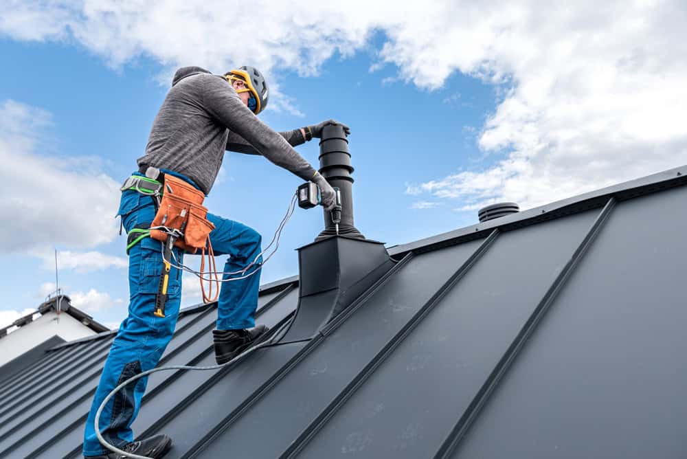 A worker in safety gear installs chimney caps on a black metal roof. He is using tools and is secured with a harness, ensuring the job is done safely and efficiently. The sky is partly cloudy.