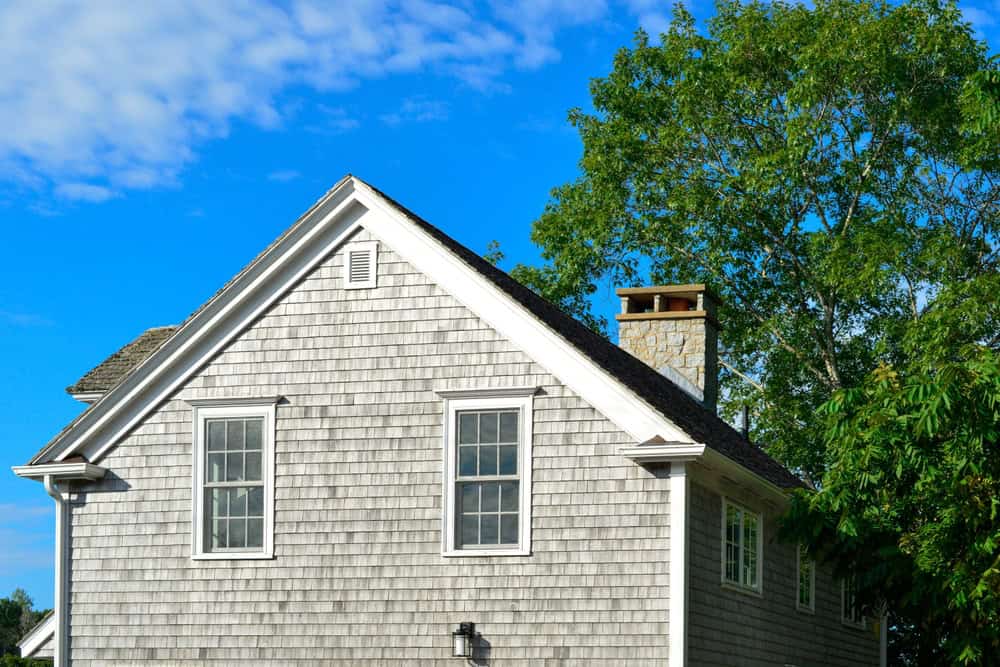 A gray shingled house with two windows on the upper level stands proudly against a clear blue sky. Showcasing expert Chimney Repair, the stone chimney complements the home's charm, while a tall, leafy tree graces the right side.