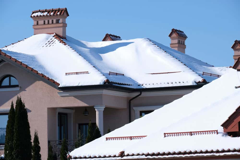 Snow-covered rooftops of a beige house with multiple chimney caps and gabled roofs stand under a clear blue sky. Tall evergreen shrubs line the house, adding contrast to the snowy scene.