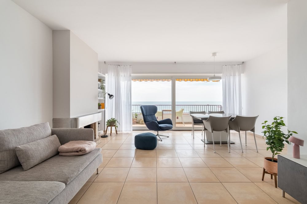 A modern living room features a gray sofa, blue armchair, and small round table. White sheer curtains cover large windows with an ocean view. Neutral tones and minimalist decor create an airy atmosphere, while a recently serviced fireplace by the Chimney Sweep adds warmth and charm.