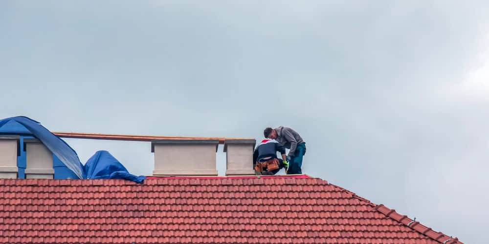 Two workers on a red tiled roof are busy with chimney repair tasks, one equipped with a tool belt. A blue tarp partially covers the chimney, hinting at ongoing work by the skilled team under the overcast sky.