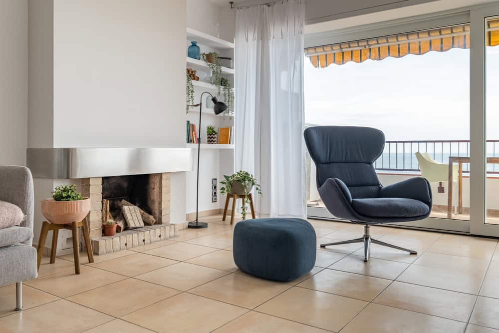 A modern living room features a sleek armchair and ottoman near the fireplace, recently serviced by a professional chimney sweep. A large window with sheer curtains offers ocean views and balcony seating, while shelves with books and plants add a cozy touch.