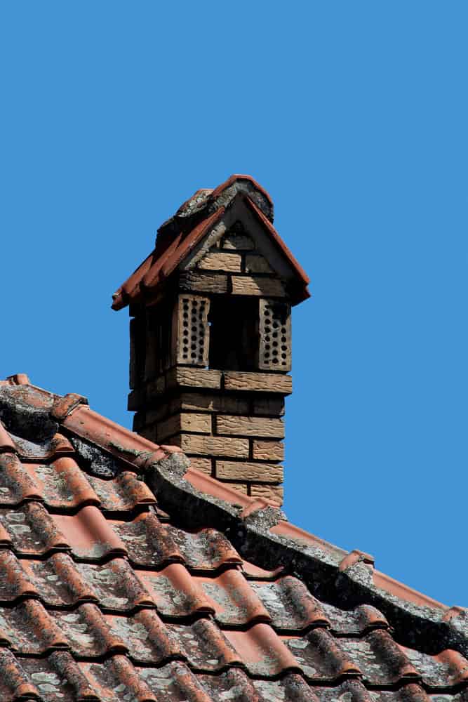 A red-tiled rooftop with a rustic brick chimney against a clear blue sky. The chimney, showcasing decorative brickwork and a small triangular roof, harmonizes with the aged appearance of the rooftop tiles—a testament to expert craftsmanship often admired by a seasoned chimney sweep.