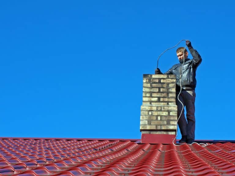 A person in a black jacket stands on a red corrugated metal roof, inspecting a brick chimney with a flexible wire under a clear blue sky. They're focused on chimney cleaning to ensure everything is spotless and safe.