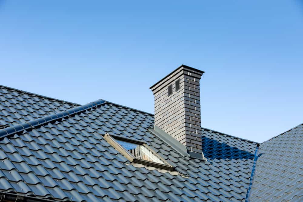 A close-up view of a house roof with dark blue tiles and a brick chimney cap under a clear blue sky. There is also a small attic window visible on the roof, showcasing the expertise of a chimney company.