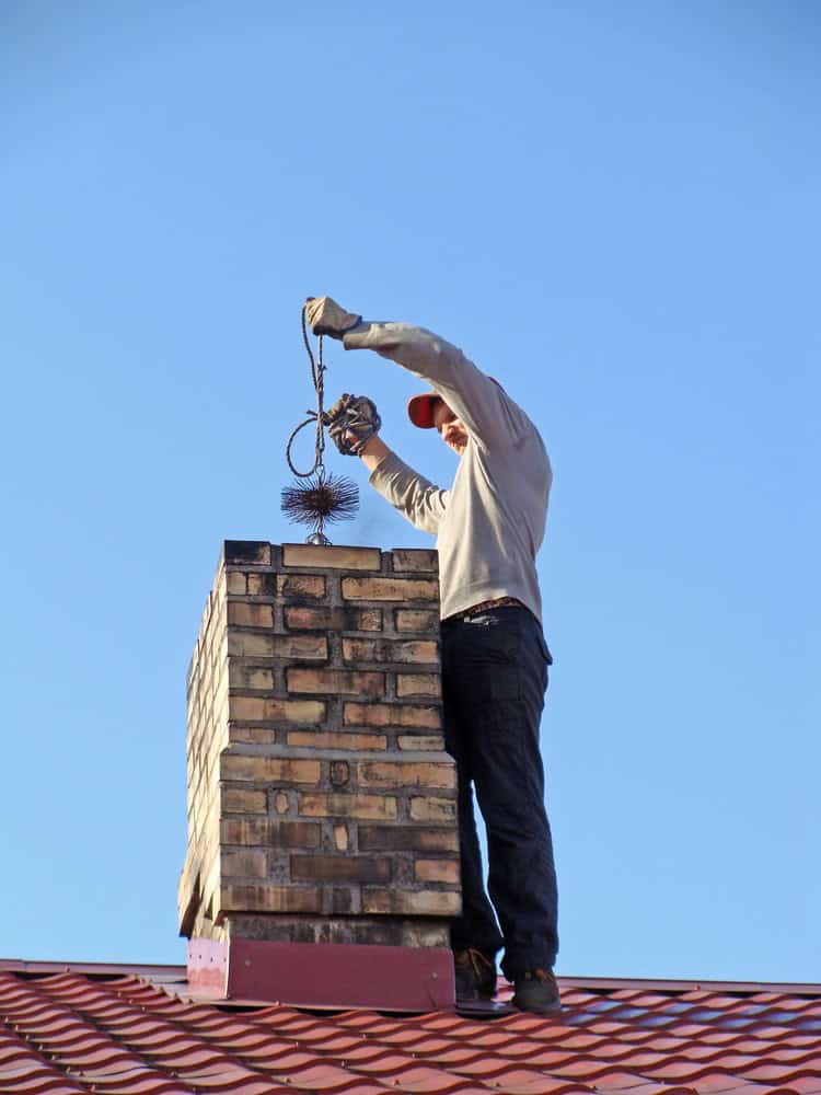 A chimney sweep wearing a hat and gloves cleans a brick chimney on a red metal roof. Using a brush attached to a cable, the worker ensures everything is spotless under the clear blue sky, showcasing the precision of their chimney company expertise.