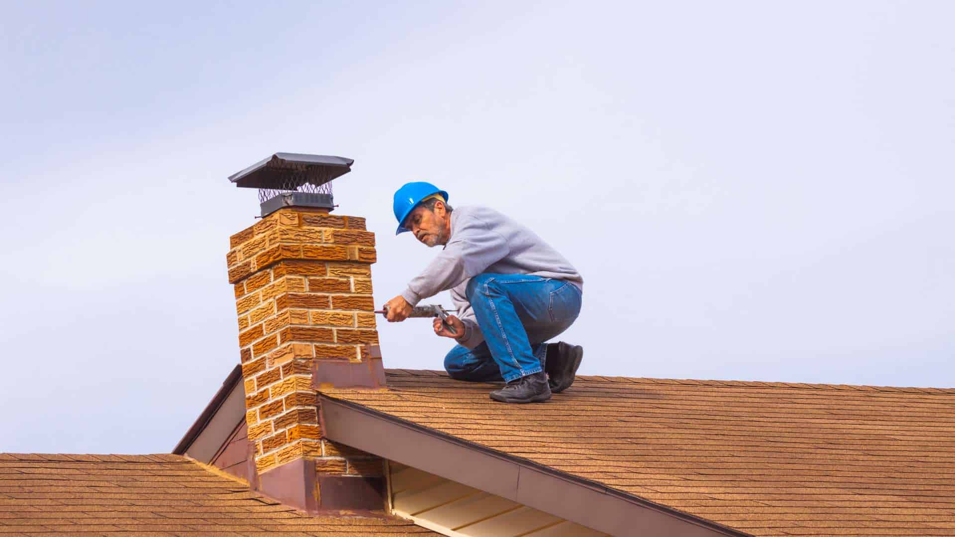 A person wearing a blue hard hat and gray sweater is crouching on a brown shingle roof. They are expertly handling chimney repair with swift precision, using a tool to work on the brick chimney, all set against a clear sky backdrop.