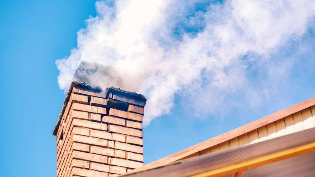 A brick chimney, recently serviced by a chimney sweep, emits white smoke against a clear blue sky, perched above a building with a sloped roof.