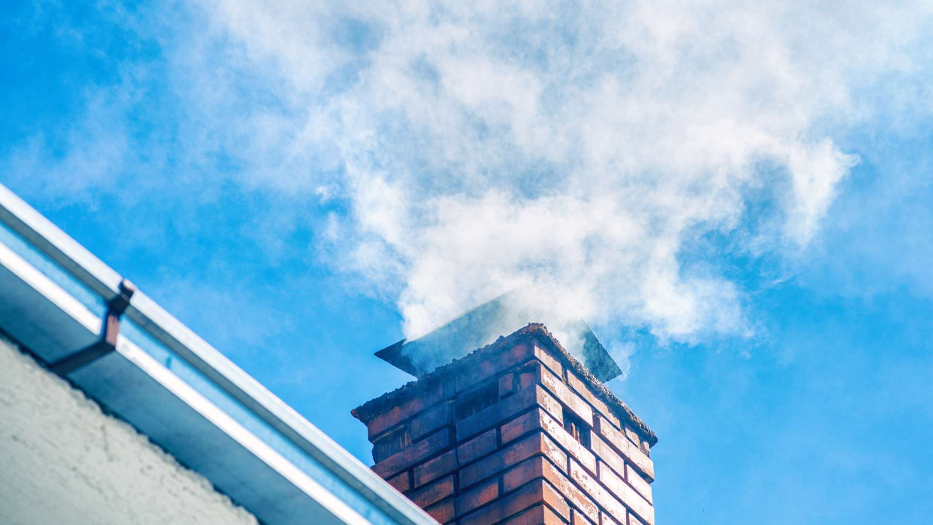 A brick chimney from a renowned chimney company emits a large plume of white smoke against a clear blue sky. A section of a sloped roof with gutters is visible on the left side of the image, showcasing flawless maintenance.