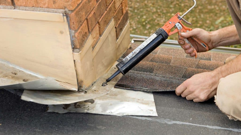 A person using a caulking gun to seal the base of a brick chimney on a shingled roof. Nearby, tools and patches for repairing roofing are scattered around, with materials ready for installing chimney caps.