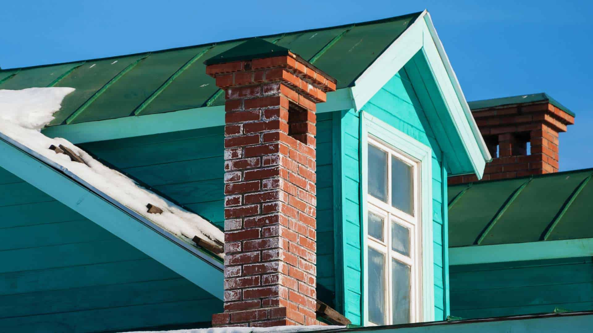 A vibrant turquoise wooden house with a green metal roof is showcased. A brick chimney, complete with a shiny chimney cap and remnants of snow, protrudes from the roof. A clear blue sky serves as the picturesque backdrop.