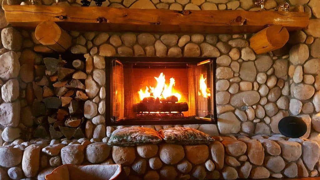 A cozy stone fireplace with a bright fire burning inside. Logs are neatly stacked to the left, and two patterned cushions rest on the hearth. The wooden mantel holds small decorative items, while a reliable chimney cap ensures safety and warmth all season long.