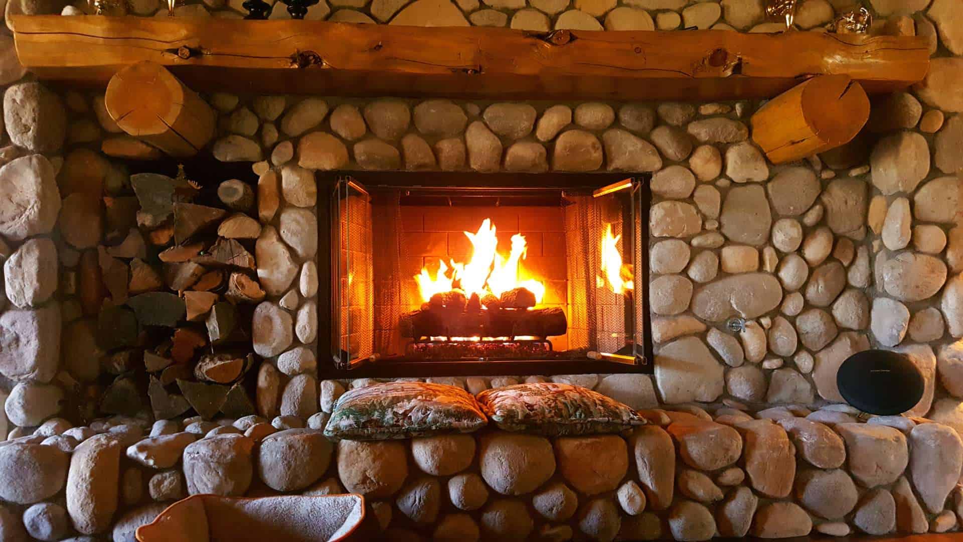 A cozy stone fireplace with a bright fire burning inside. Logs are neatly stacked to the left, and two patterned cushions rest on the hearth. The wooden mantel holds small decorative items, while a reliable chimney cap ensures safety and warmth all season long.