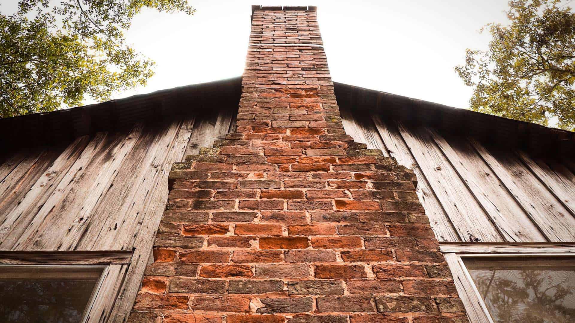 A low-angle view of a rustic brick chimney on a wooden house, framed against a pale sky. The weathered wooden planks and surrounding green foliage add to the scene's vintage charm, while hints of recent chimney cleaning ensure safety amidst its natural elegance.