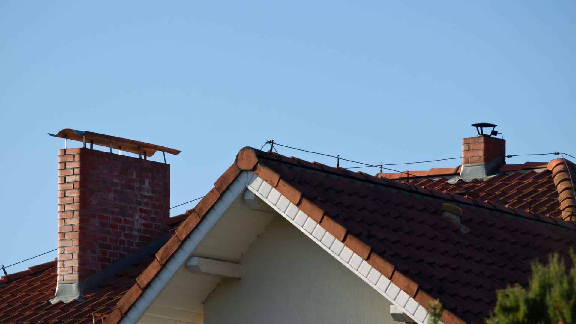 A rooftop scene with red tiles, two chimneys, and a clear blue sky. The brick chimneys stand tall; one has a metal cover hinting at recent chimney repair. The roof edges are white, with sunlight casting shadows on the tiles.