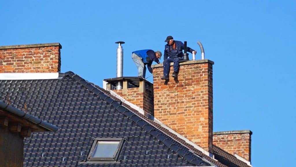 Two workers from a chimney repair company, clad in safety gear, are inspecting chimneys on the roof of a brick building. The roof, adorned with dark tiles and featuring a small skylight, sits under the clear blue sky.